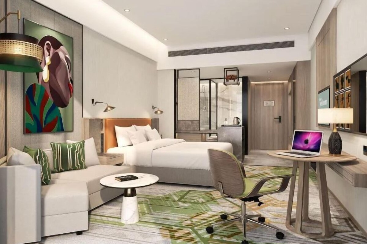 Una habitación de hotel moderna con una cama, un sofá, un escritorio con una computadora portátil y un cuadro grande en la pared.