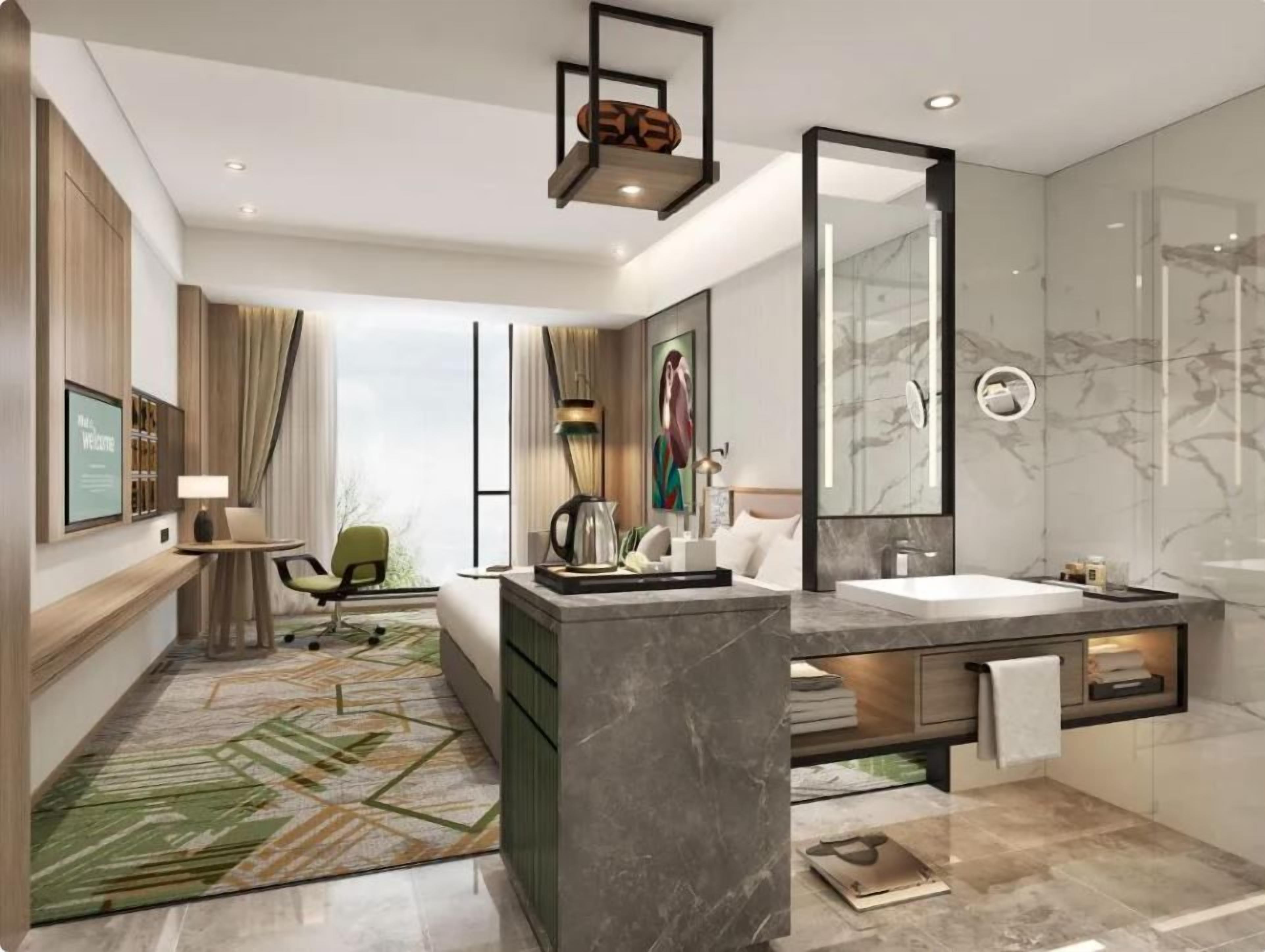 A modern hotel room with a large bed, a sitting area with a desk and chair, a large mirror, a bathroom with a marble sink and shower, and a green patterned rug.