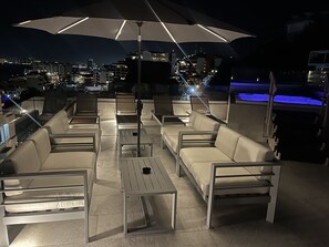 Un espace extérieur aménagé avec des sièges, un grand parasol, plusieurs chaises et une piscine en arrière-plan.
