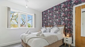 A bedroom with a large window, a floral wallpaper, a bed with pillows and a blanket, and a bedside table with a lamp. - 3 Bedroom Home Sleeps 7 OR 8 Free Parking NR Solihull Edgbaston and City Centre (Birmingham)