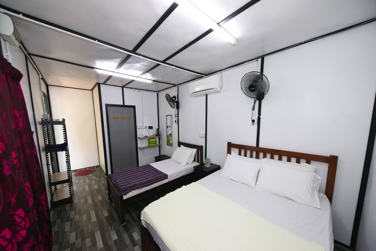 Habitación de hotel compacta con una cama de madera, una mesita de noche pequeña, aire acondicionado montado en la pared y un ventilador de techo.