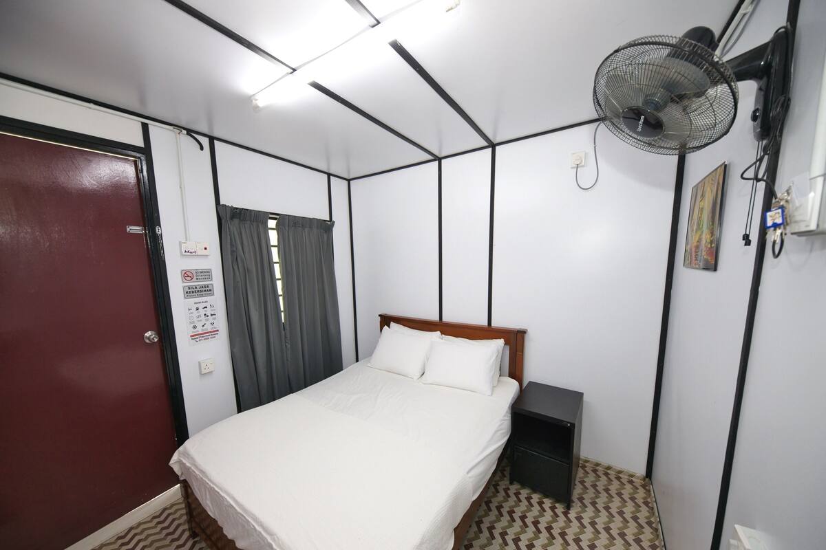 Una habitación de hotel con cama, mesita de noche, ventilador de pared y una puerta.