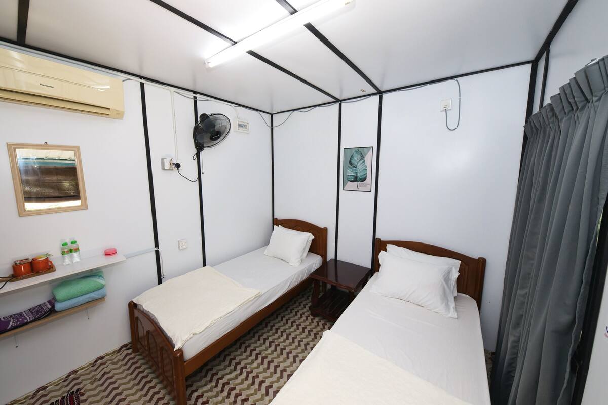 Una habitación pequeña y limpia con dos camas individuales, un ventilador, un estante con artículos de aseo y una ventana con persianas.