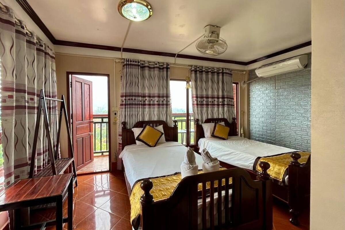 A hotel room with two single beds, a wooden table, a bench, a ceiling fan, and a window with curtains.