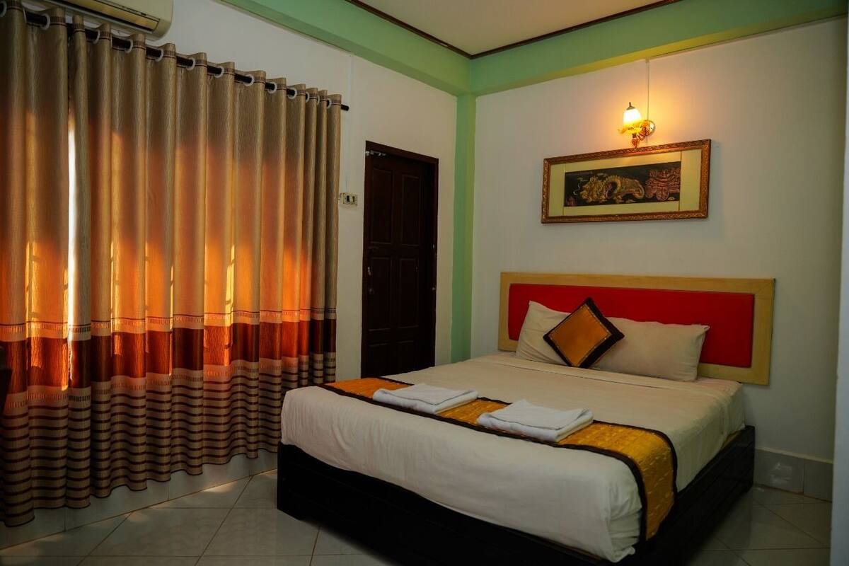 A hotel room with a bed, curtains, a painting, and a wall-mounted light.