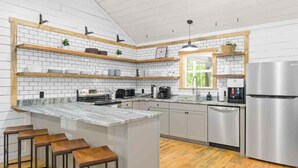 A modern kitchen with white subway tiles, wooden shelves, a marble island, bar stools, stainless steel appliances, and a window with a view.