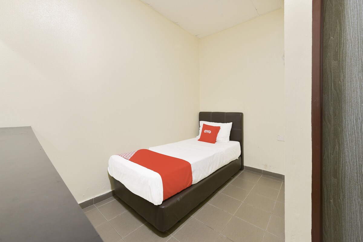 A small, single-bed room with a dark headboard, white bedding, and a red accent pillow.