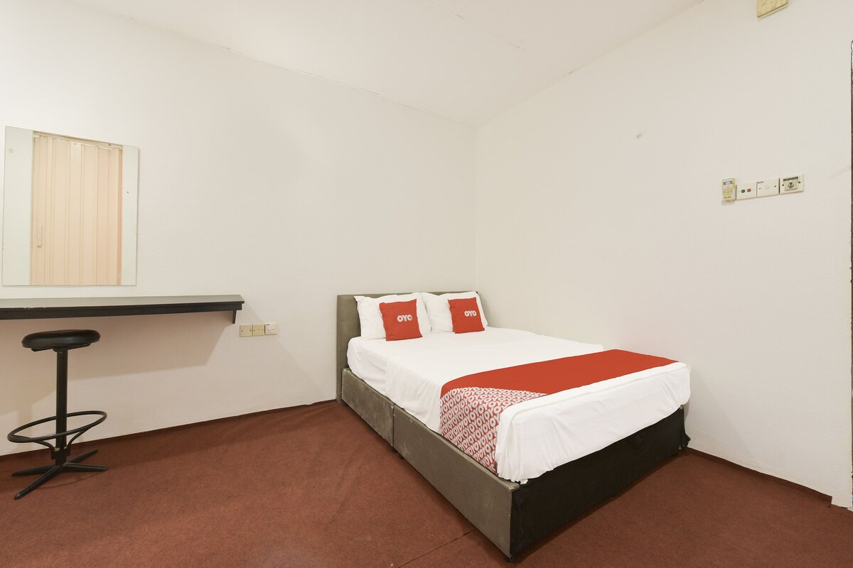 A single bed with white bedding and red pillows, a small desk with a chair, and a wall-mounted light switch.