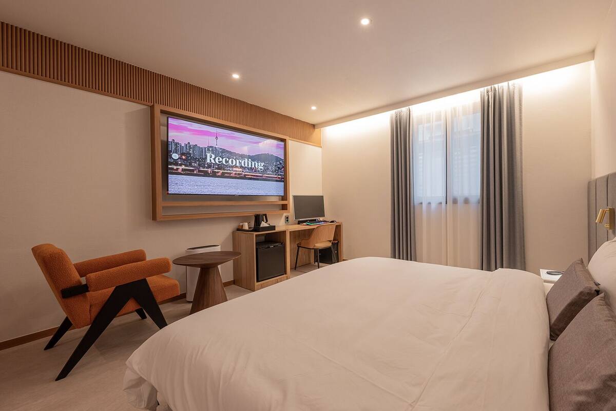 A hotel room with a large bed, a desk with a computer, a TV mounted on the wall, and a seating area with a chair.