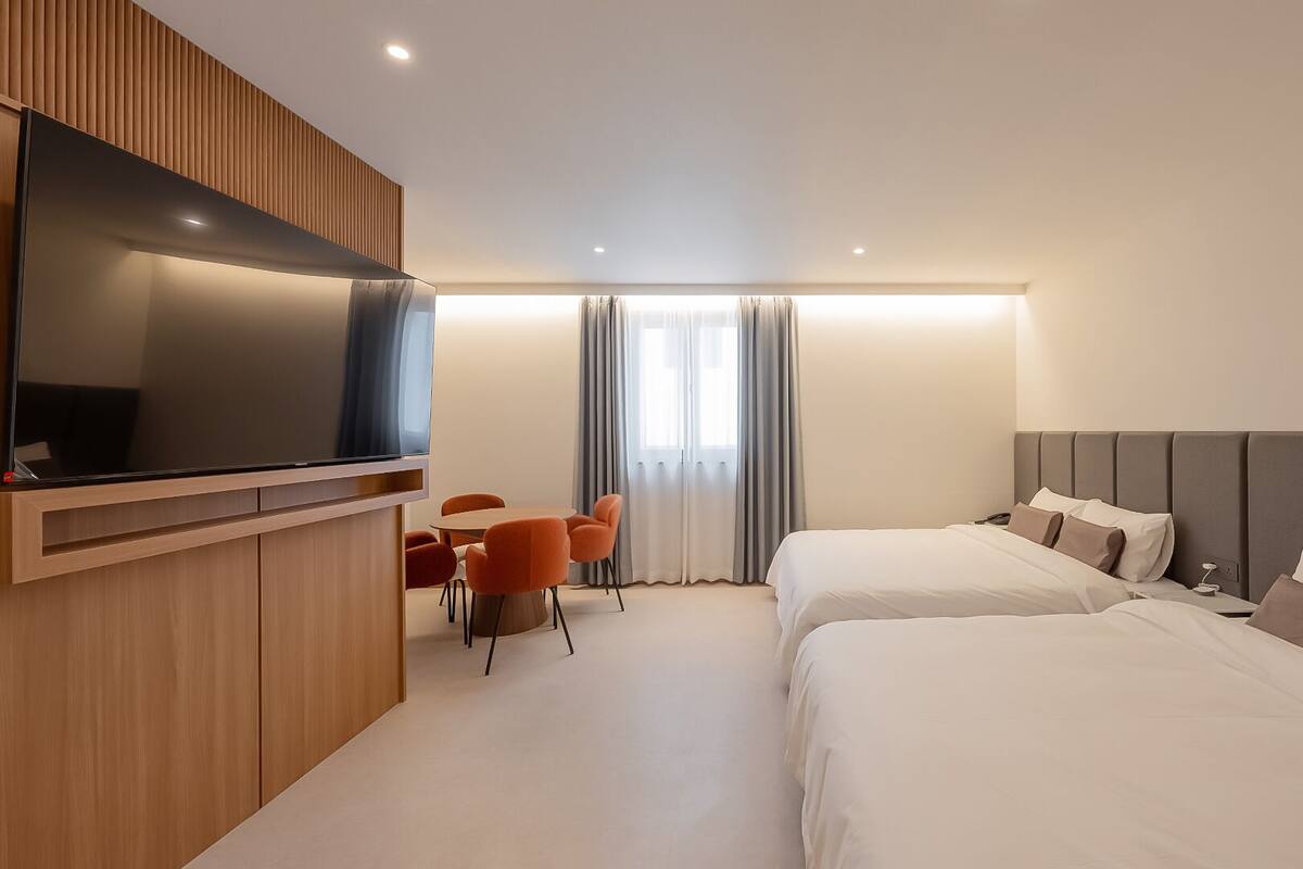 A hotel room with two beds, a TV mounted on a wooden panel, a small table with chairs, and a window with curtains.