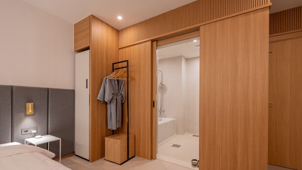 A modern hotel room with a bed, bedside table, and a wooden wardrobe with a sliding door leading to a shower area. - H Avenue Munhyeon (Busan)