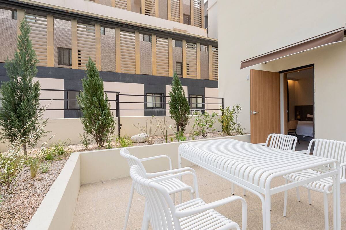 A modern outdoor patio with a white table and chairs, surrounded by plants and a view of a building with a geometric facade.