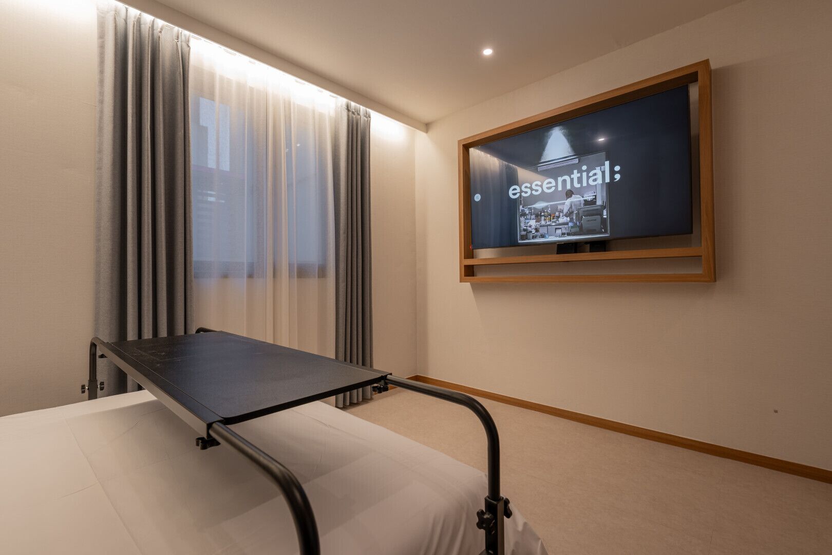 A hotel room with a bed, a window with curtains, and a wall-mounted television displaying the word 'essential'.