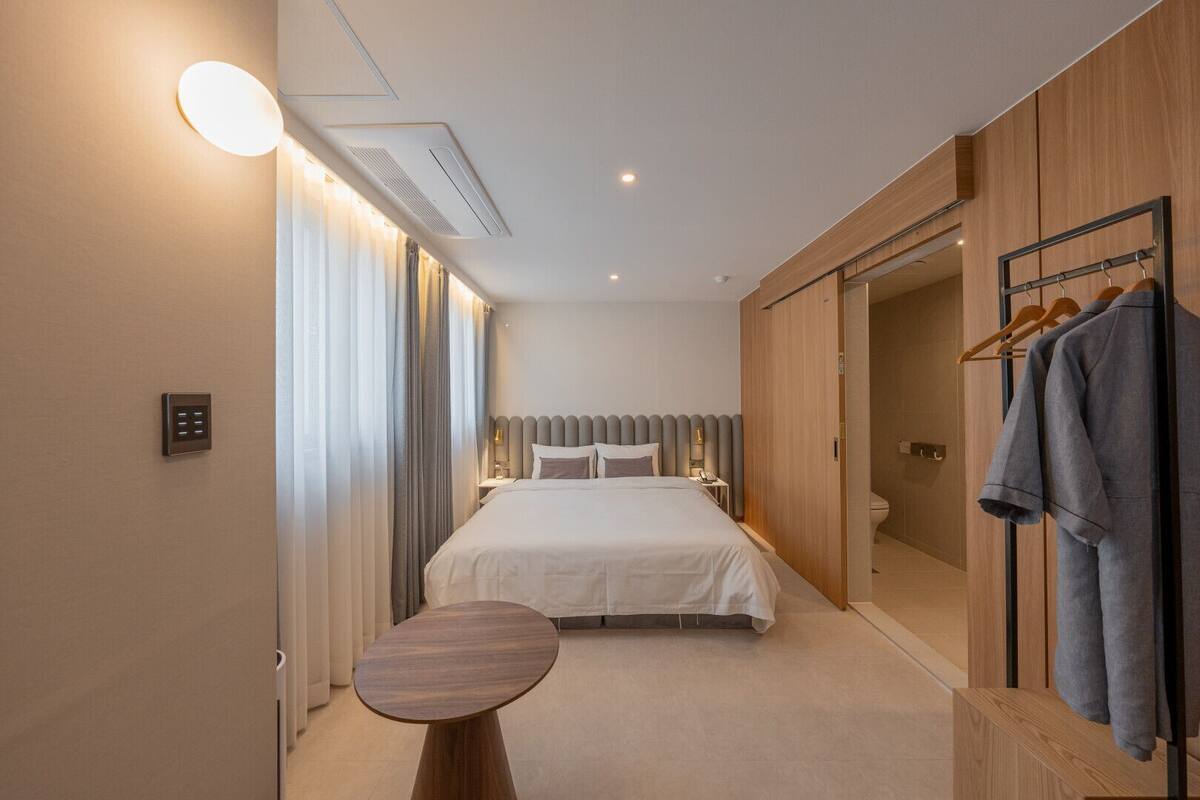 A modern hotel room with a large bed, bedside tables, a small round table, and a wardrobe with hanging clothes.
