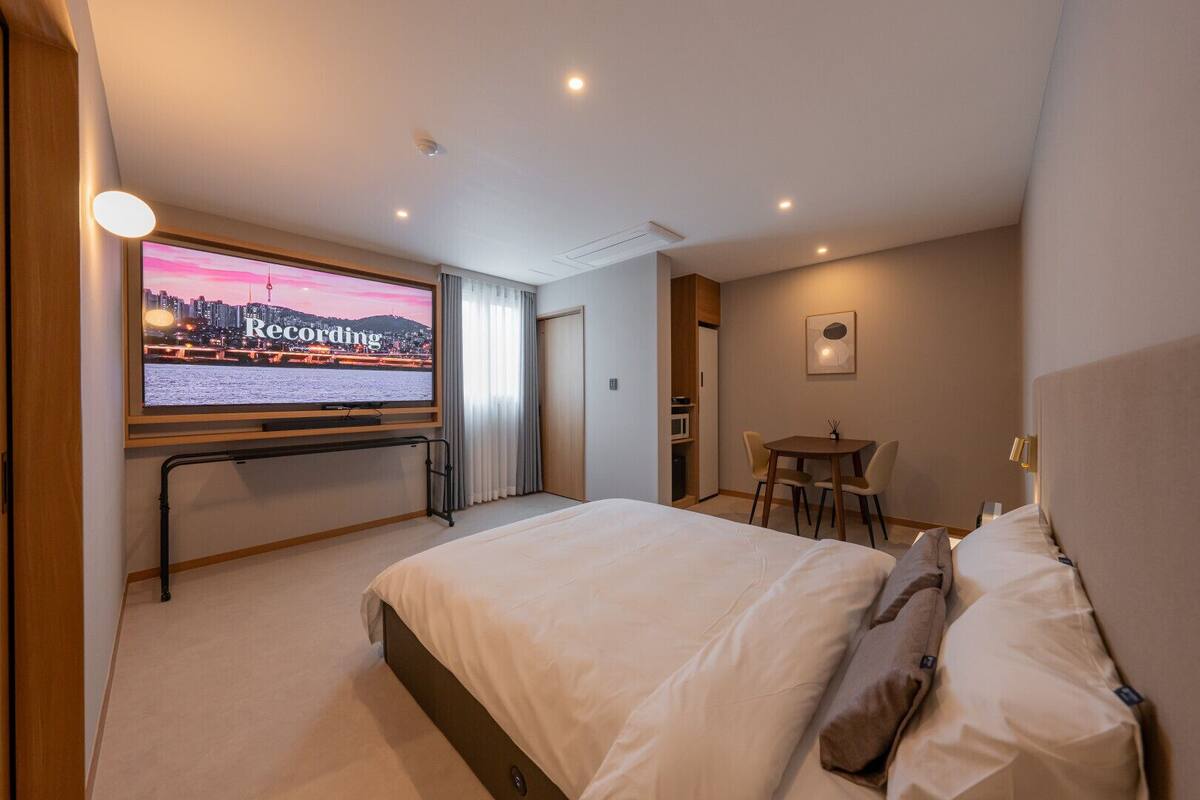 A hotel room with a large bed, a desk with chairs, a television displaying a cityscape, and a door leading to a bathroom.