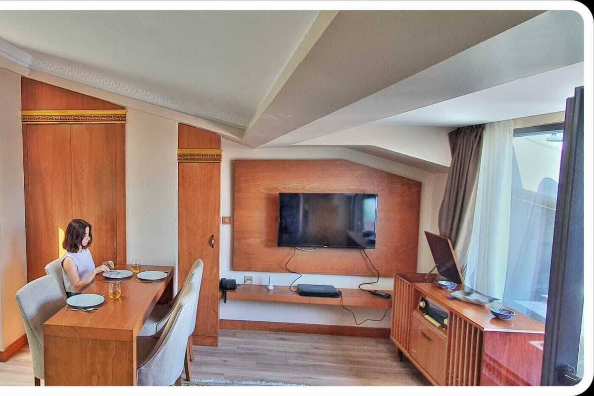 A modern hotel room with a dining area, a flat-screen TV mounted on the wall, and a large window providing a view.