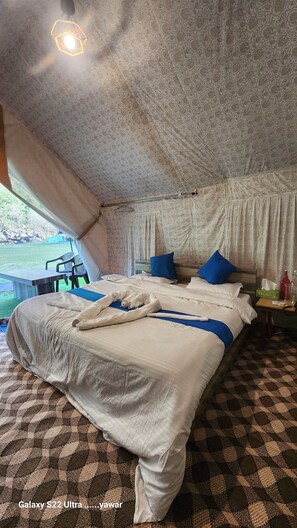 A tent-style room with a large bed, two blue pillows, a bedside table with a bottle and tissues, and a view of a grassy area outside. - Snowber Resorts (Gund)
