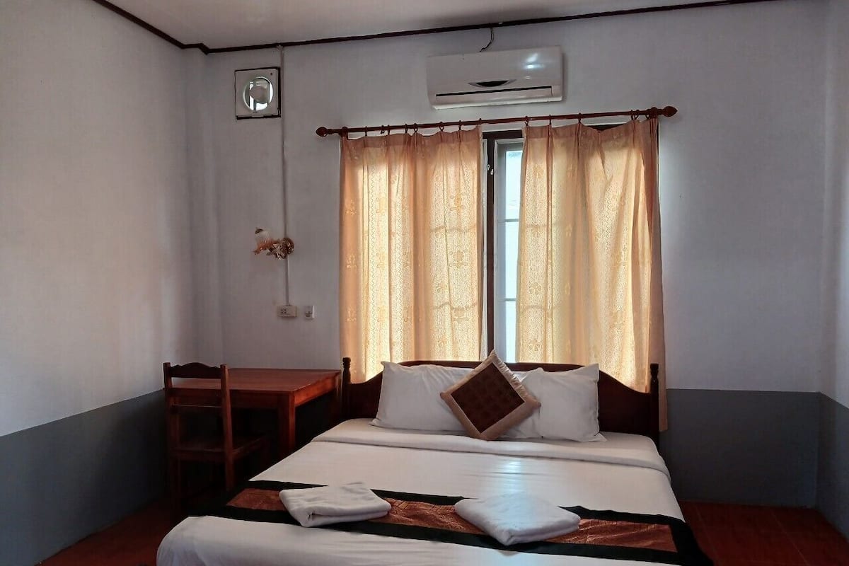 A hotel room with a single bed, a wooden desk, a window with curtains, and an air conditioning unit.