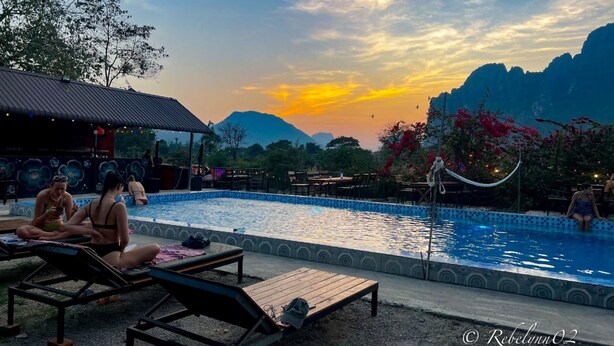 A poolside area with lounge chairs and a covered seating area, surrounded by mountains and a sunset sky. - Riverside Garden Hostel (Vang Vieng)