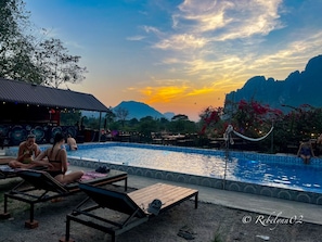A poolside area with lounge chairs and a covered seating area, surrounded by mountains and a sunset sky. - Riverside Garden Hostel (Vang Vieng)
