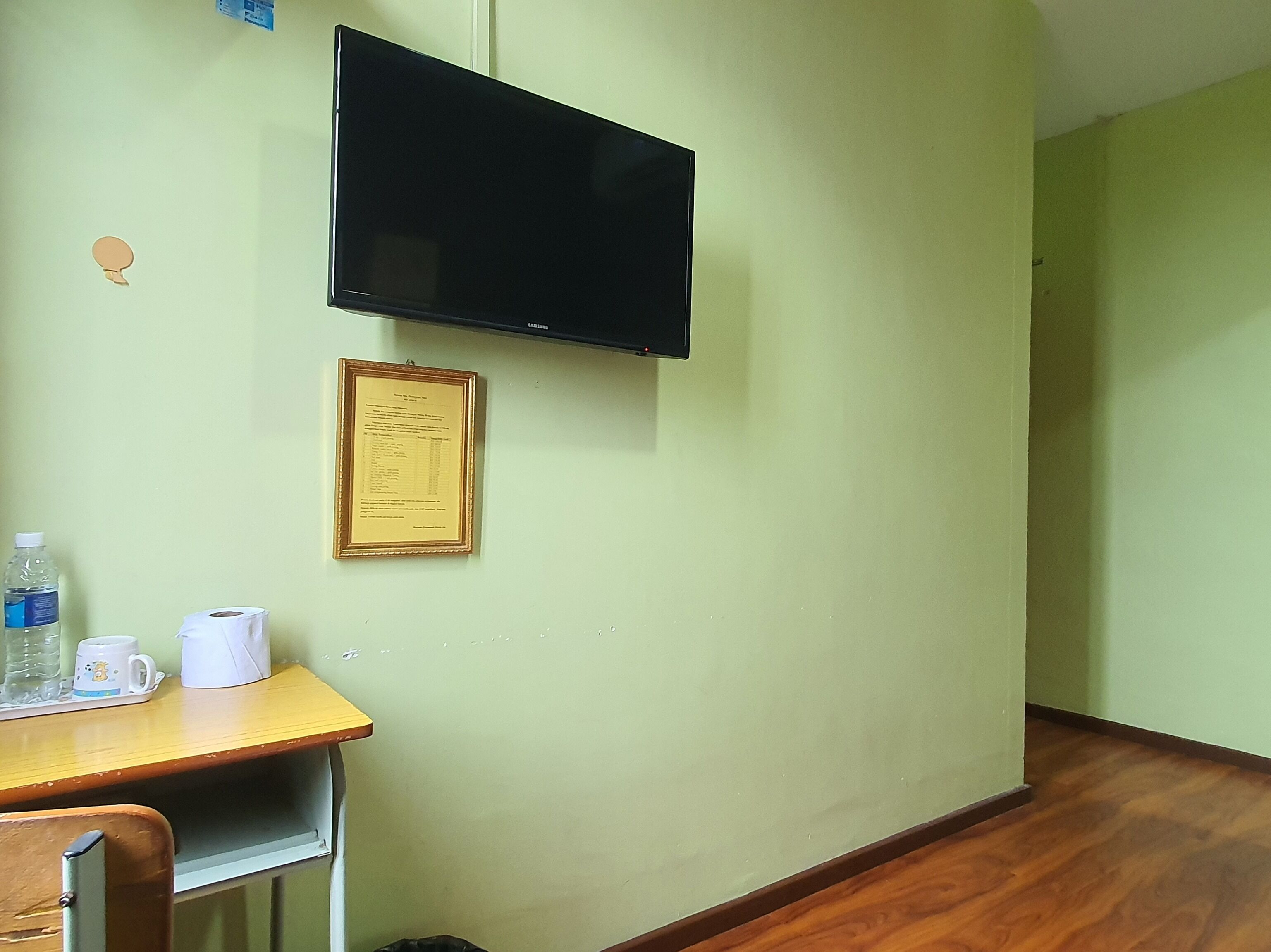 Una habitación con escritorio, televisor de pantalla plana montado en la pared, un certificado enmarcado, una botella de agua, una taza de café y un cubo de basura.