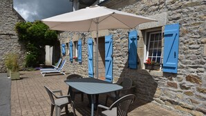 Ein Steinhäuschen mit einer Terrasse, ausgestattet mit Tisch, Stühlen und einem großen Sonnenschirm, sowie blauen Fensterläden.