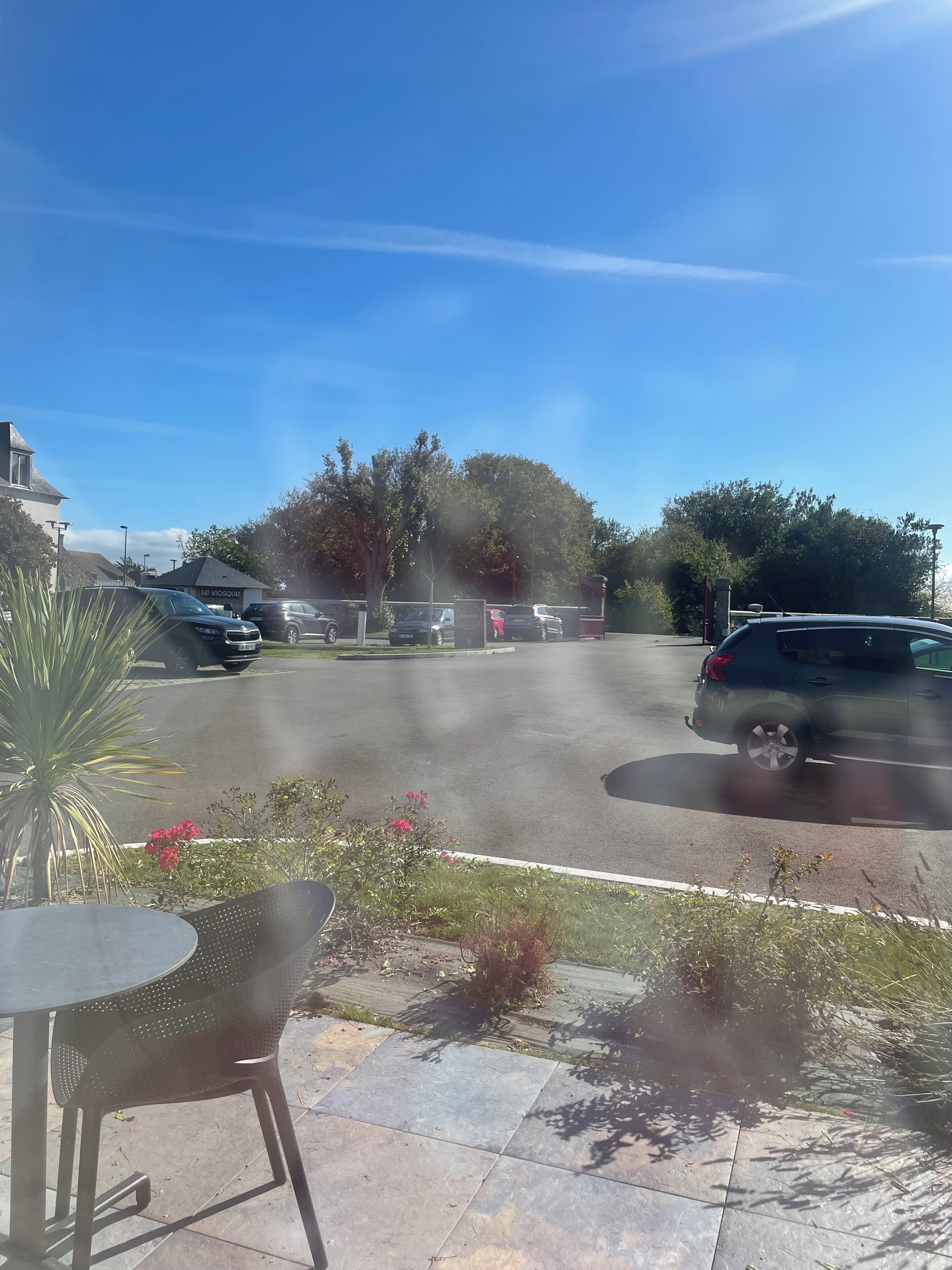 A parking lot with several cars, a building labeled "Le Kiosque," and a patio area with a table and chairs.