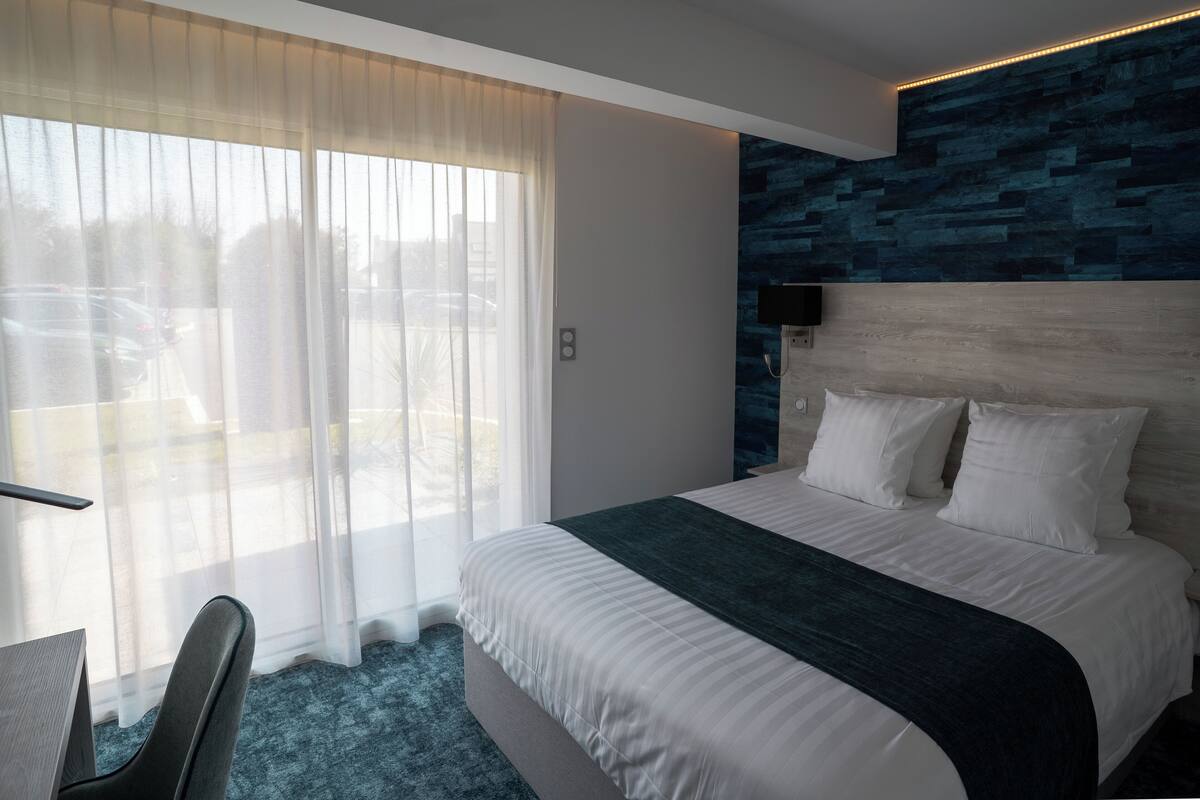 A modern hotel room with a large bed, a desk, and a view of the parking area through sheer curtains.