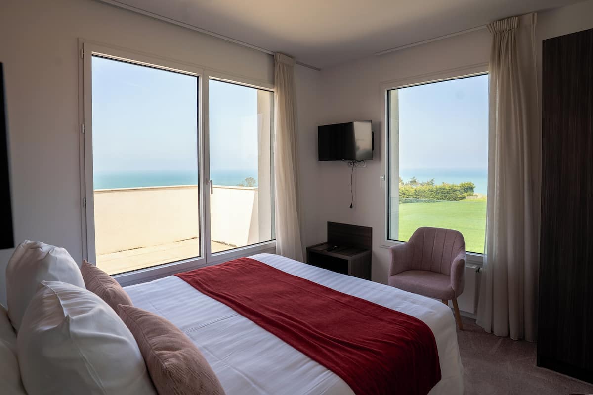 A hotel room with a large bed, a TV, a chair, and a view of the sea.