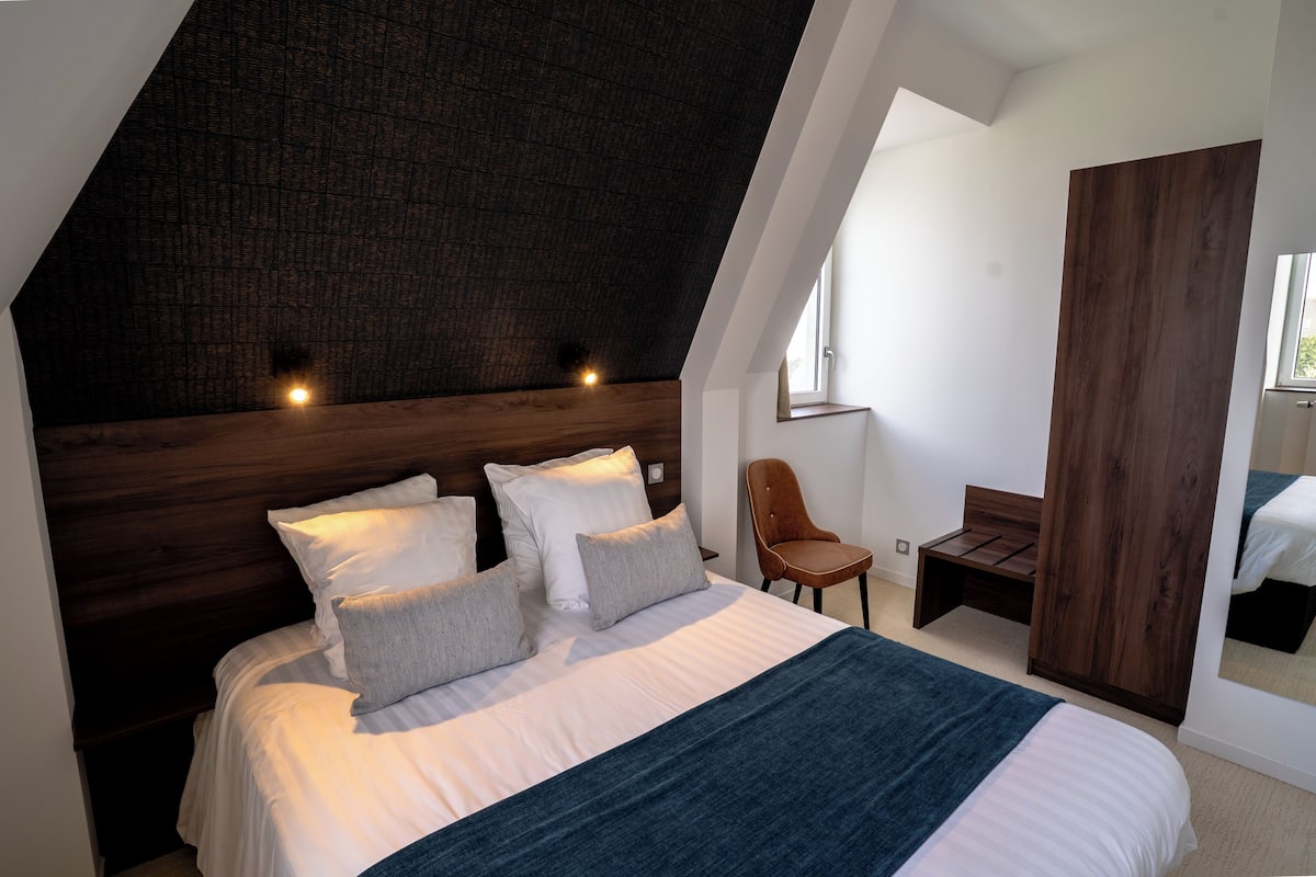 A modern bedroom with a sloped ceiling, a large bed with white linens and multiple pillows, a wooden headboard, a wooden bench, and a chair.