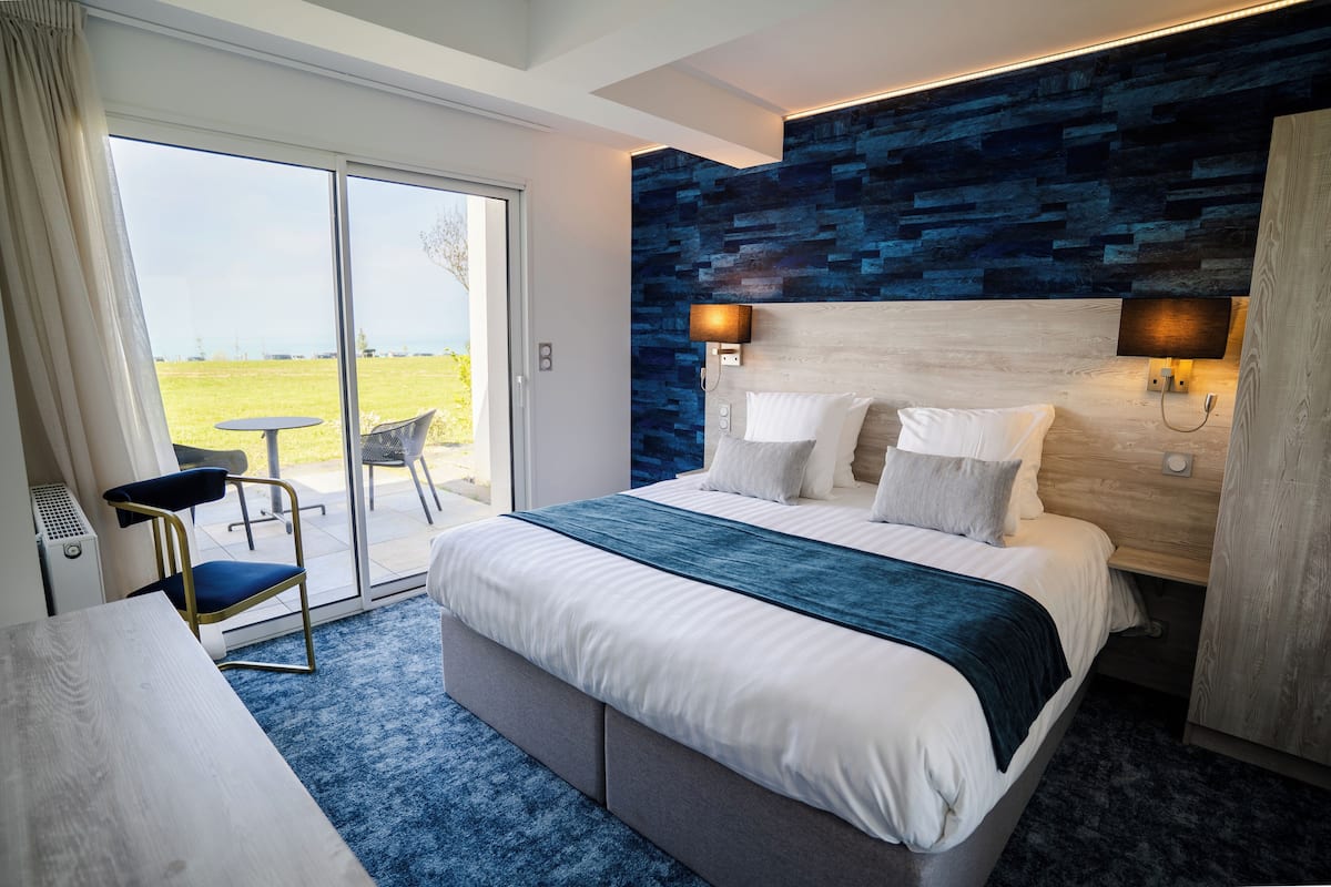A modern hotel room with a large bed, a desk, a chair, and a view of the outdoors through a sliding glass door.