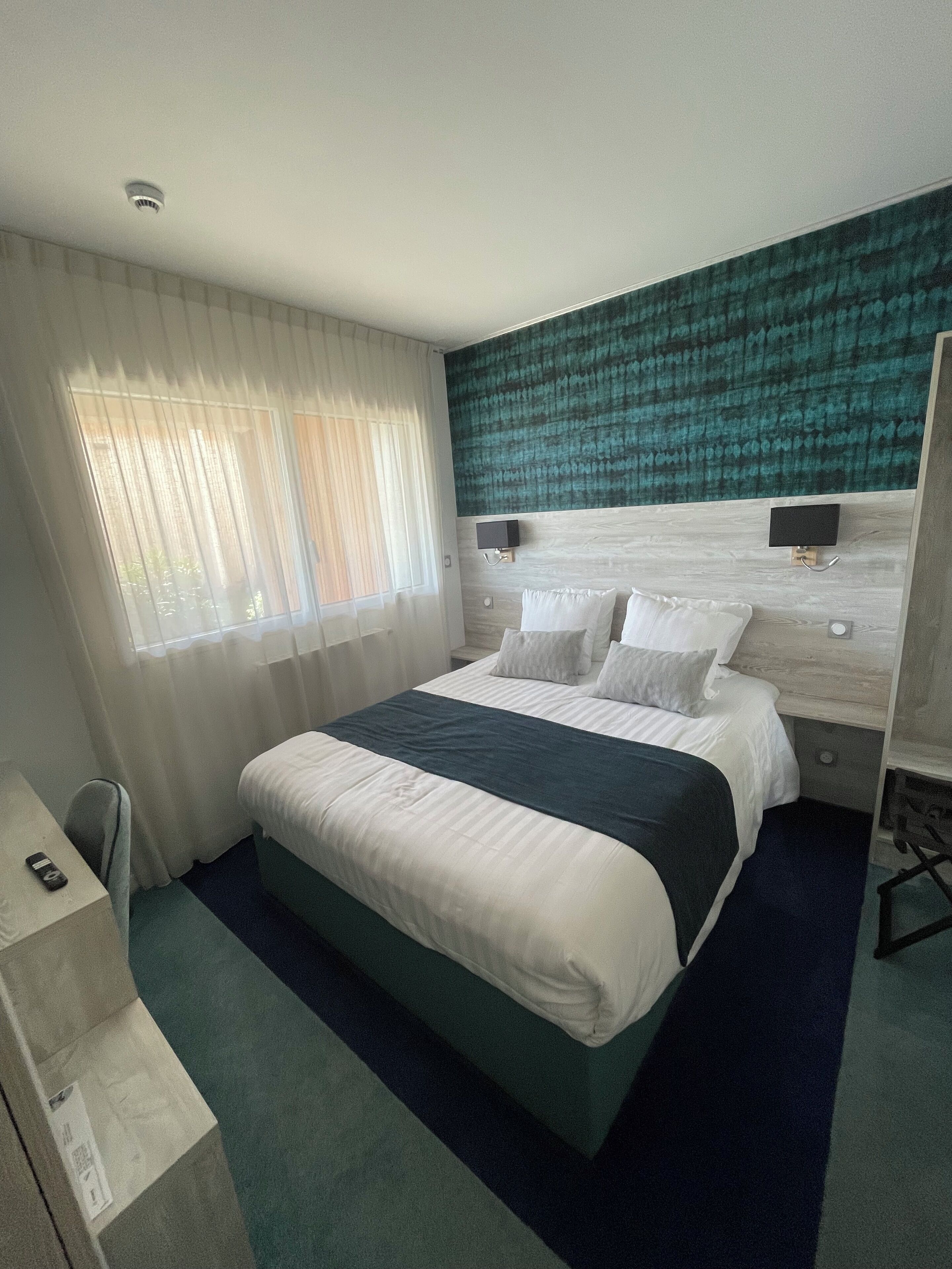 A hotel room with a large bed, a headboard with a textured design, a bedside table with a lamp, and a window with sheer curtains.