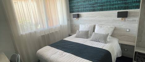 A hotel room with a large bed, a headboard with a textured design, a bedside table with a lamp, and a window with sheer curtains.
