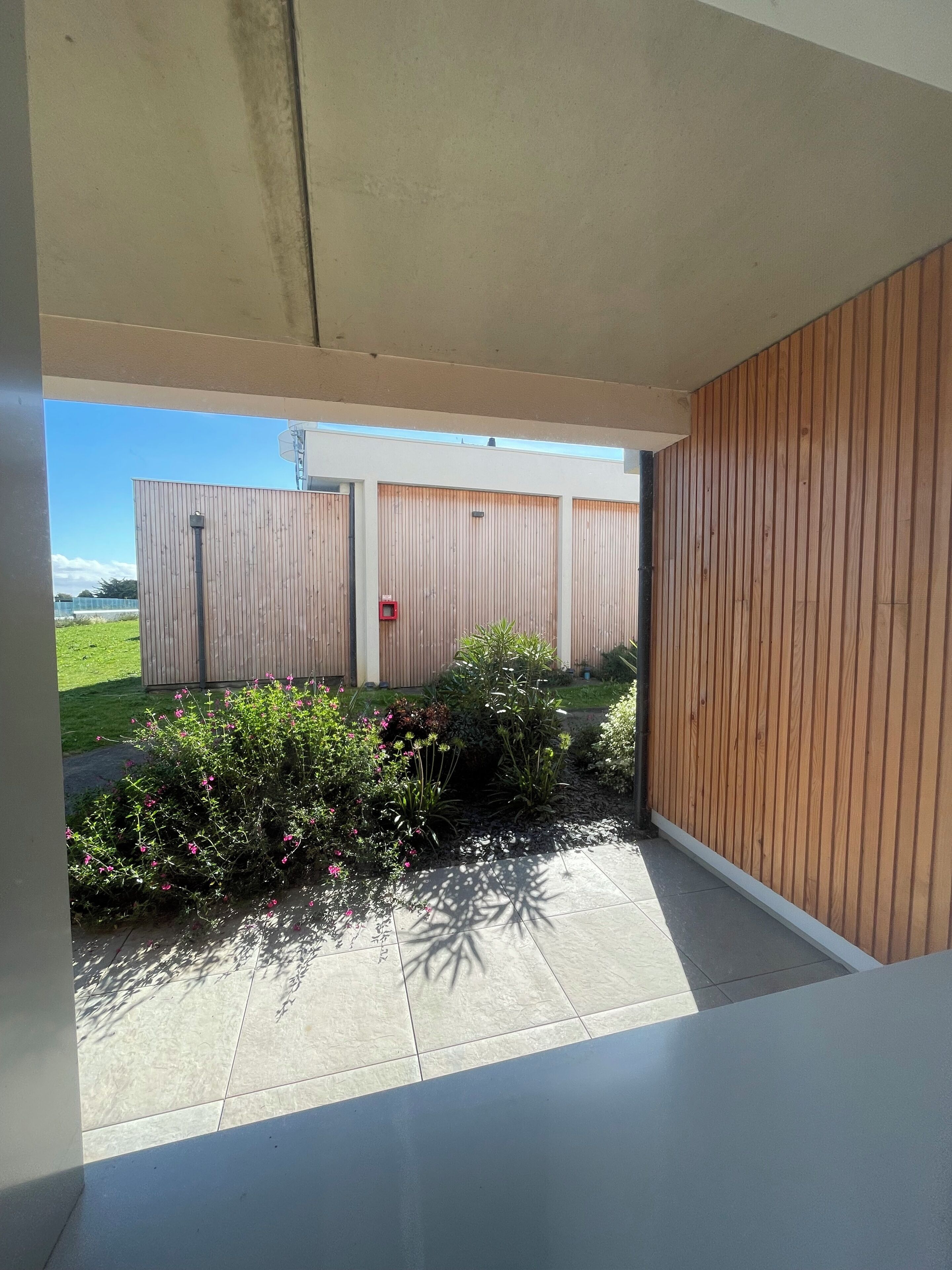 A covered outdoor area with a concrete floor, wooden panel walls, and a view of a fenced property with greenery.