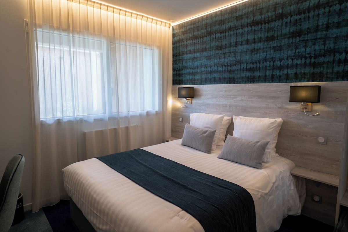 A modern hotel room with a large bed, two bedside tables with lamps, a headboard with a textured design, and a large window with sheer curtains.
