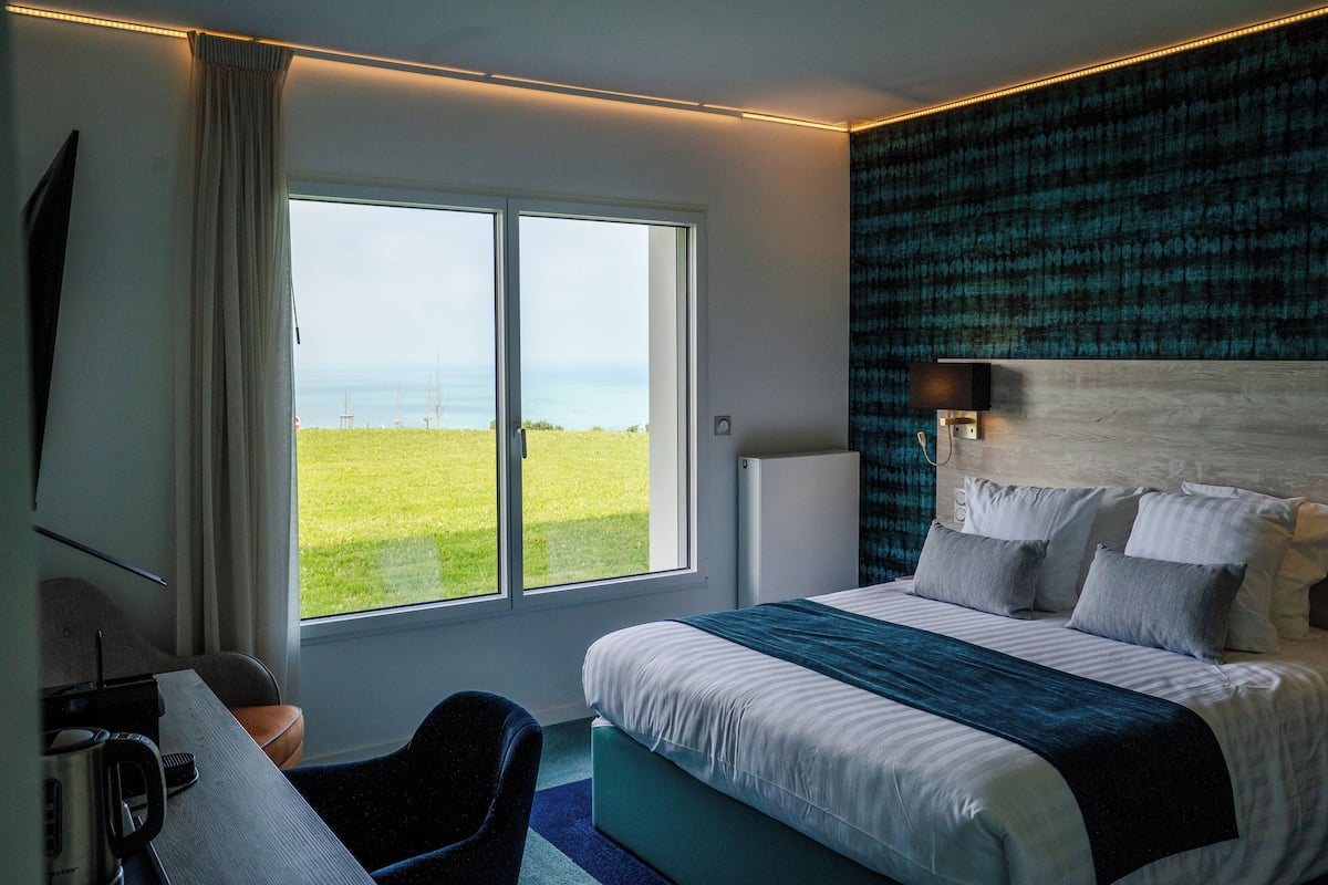 A modern hotel room with a large bed, a desk with a chair, a view of the ocean, and a patterned wall.