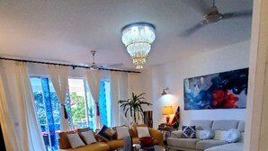 A spacious living room with a large beige sofa, a smaller sofa, and a coffee table. There is a chandelier hanging from the ceiling and a painting on the wall. - Gustavo Homes Mombasa (Mombasa)
