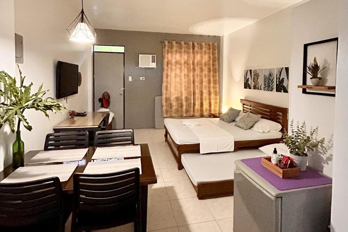Habitación de hotel compacta con cama, escritorio, mesa de comedor con sillas y kitchenette.