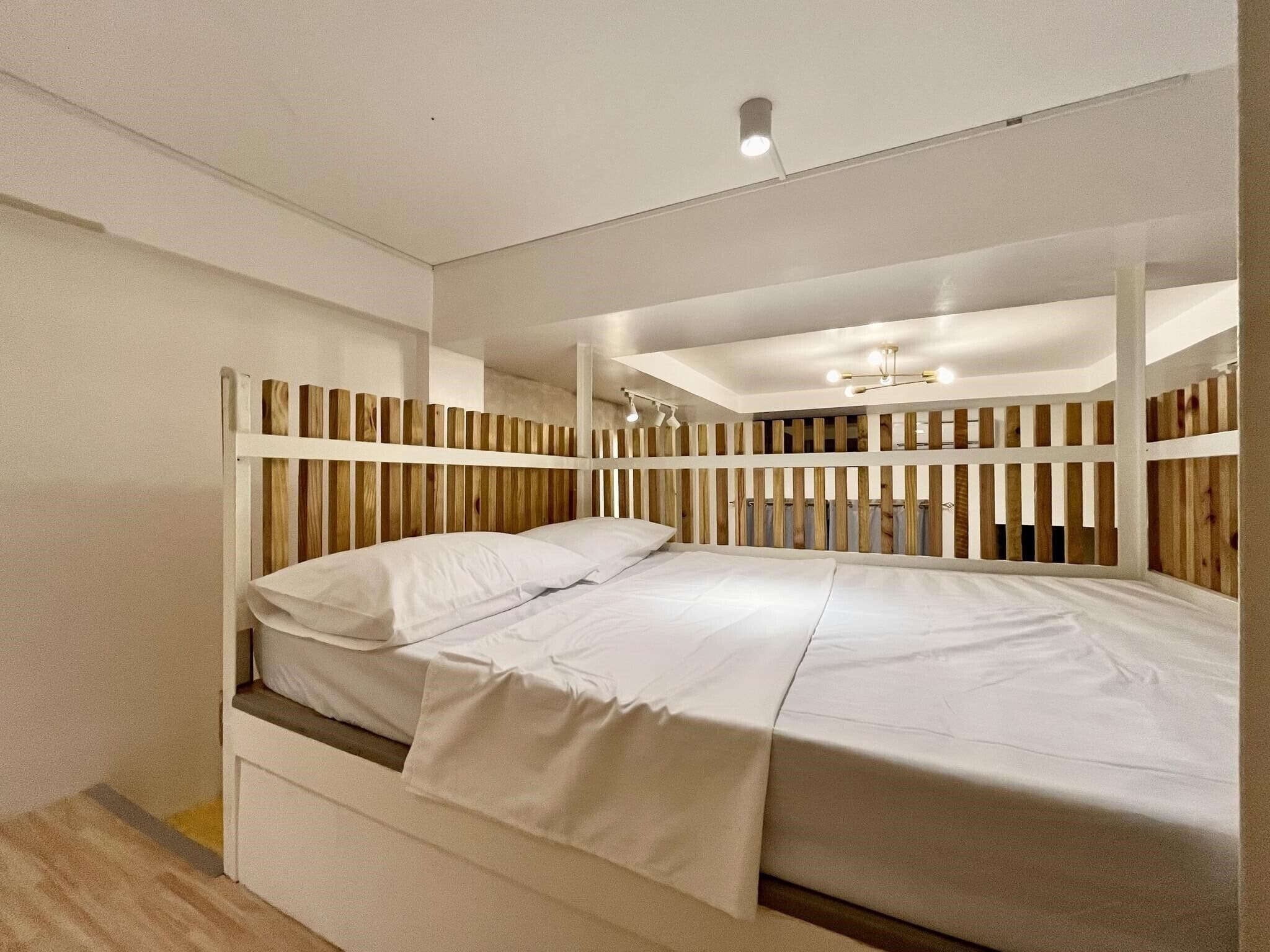 Uma cama de solteiro com cabeceira e peseira de madeira, roupa de cama branca e luminária no teto.