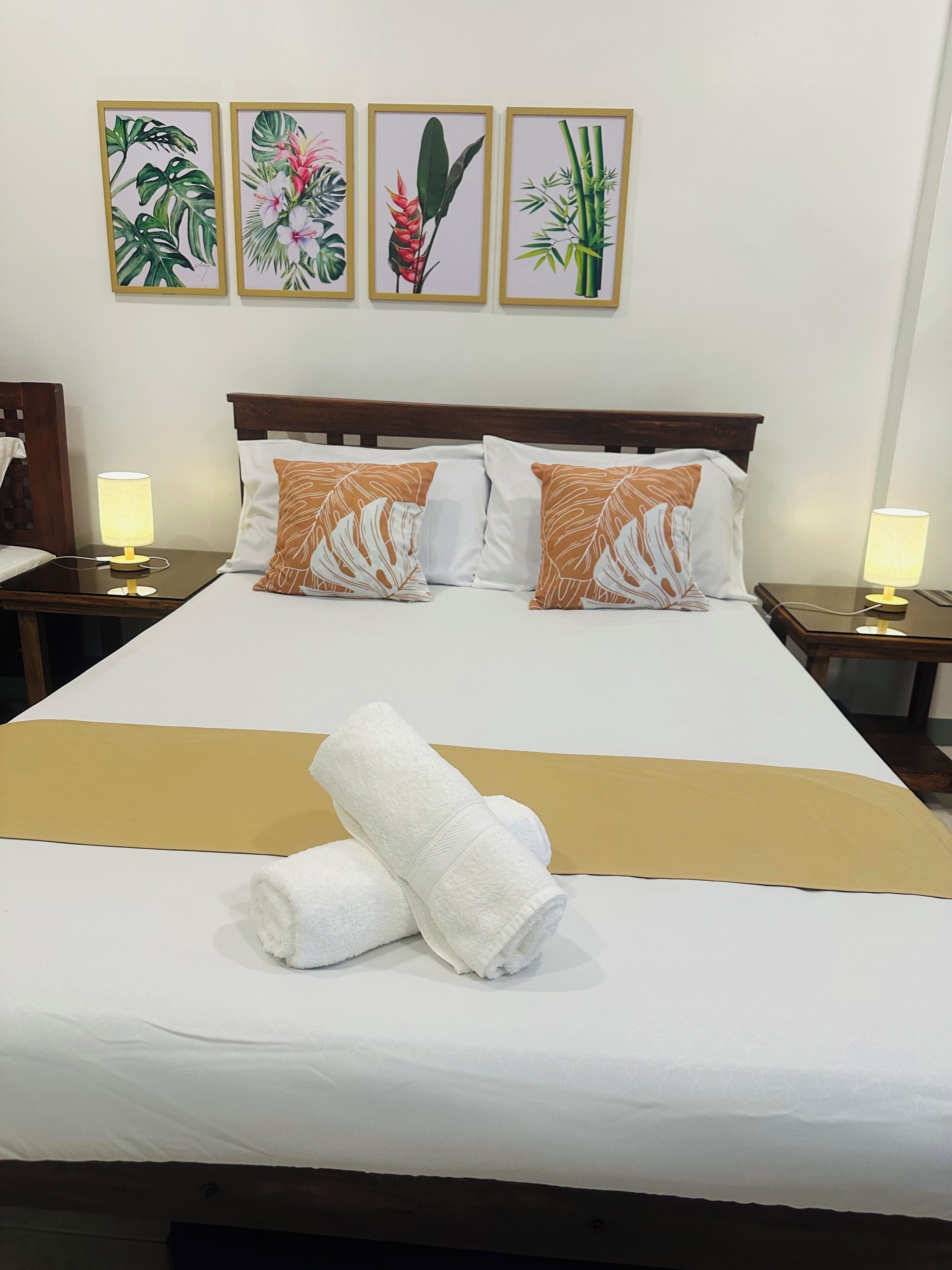 Cama bem arrumada com roupa de cama branca, duas almofadas castanhas e uma toalha dobrada. Há duas mesas de cabeceira com luminárias e quatro gravuras botânicas emolduradas na parede.