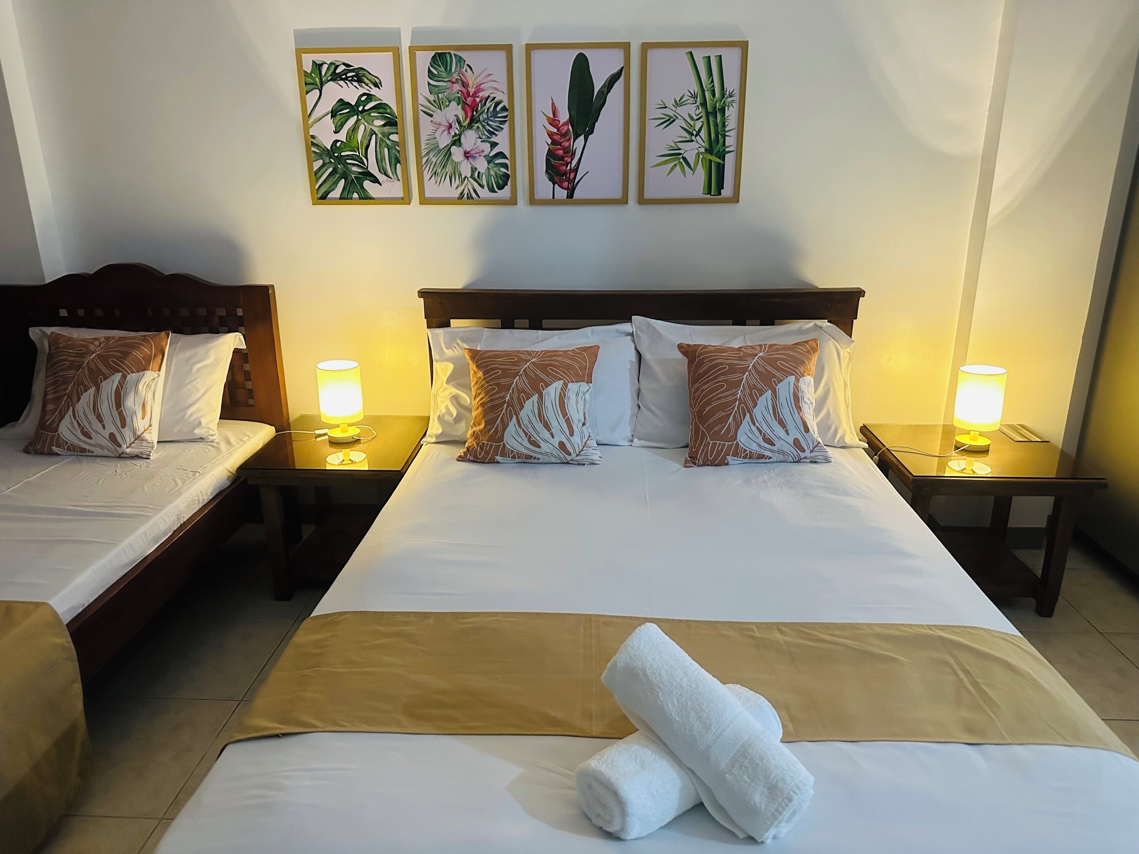 A hotel room with two beds, a large bed with a brown bedspread and two pillows, and a smaller bed with a white bedspread and two pillows. There are two bedside tables with lamps, and a wall with four framed botanical prints.