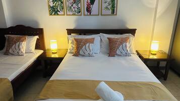 A hotel room with two beds, a large bed with a brown bedspread and two pillows, and a smaller bed with a white bedspread and two pillows. There are two bedside tables with lamps, and a wall with four framed botanical prints.