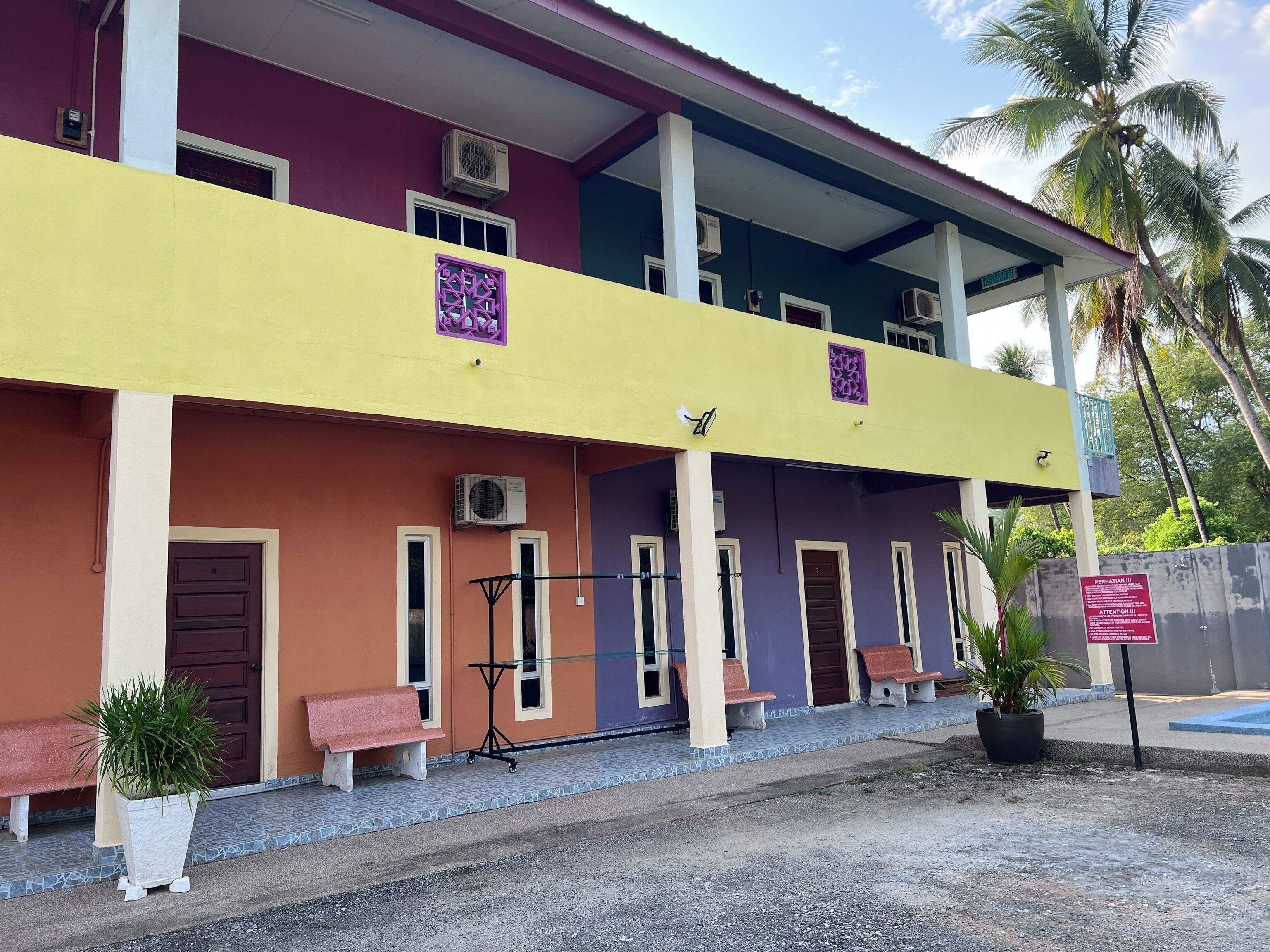 A multi-story building with a yellow and purple facade, air conditioning units, and a covered entrance area with benches and potted plants.