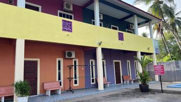 A multi-story building with a yellow and purple facade, air conditioning units, and a covered entrance area with benches and potted plants.