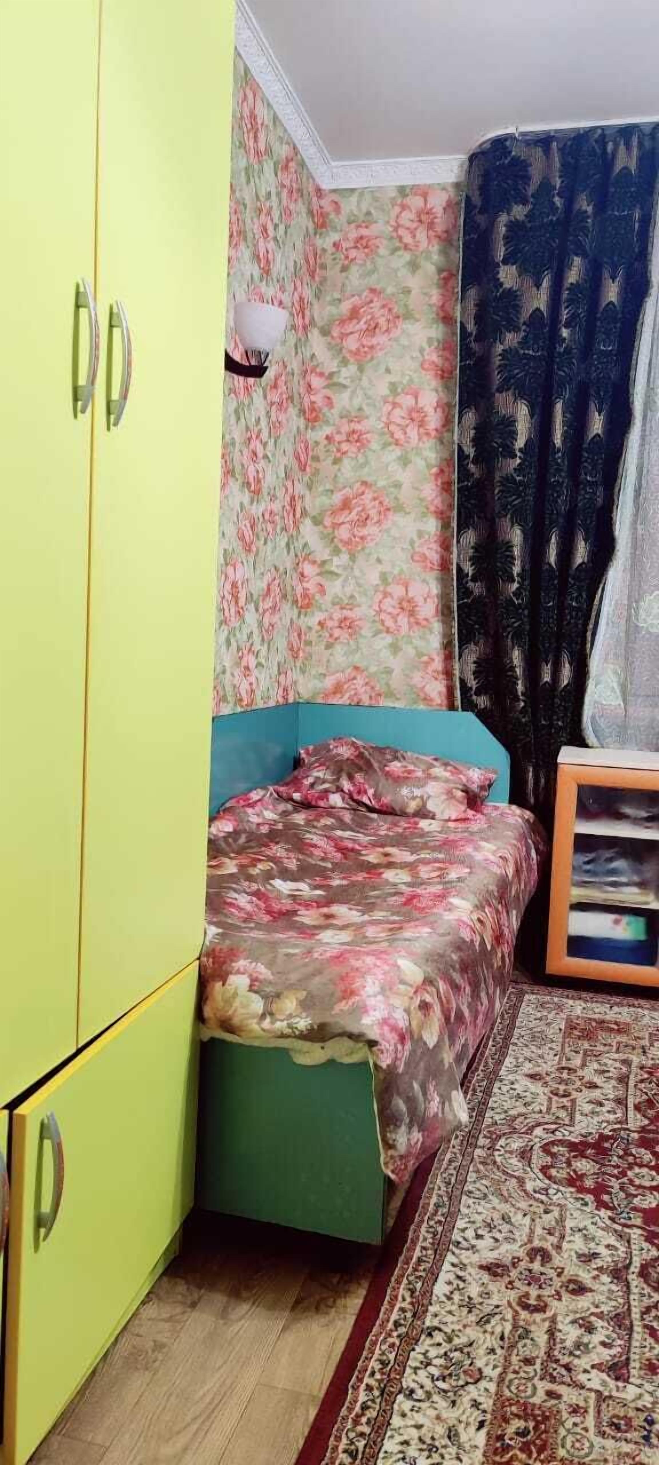 A small room with a bed, a green wardrobe, a floral-patterned wall, and a patterned rug.