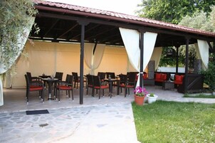 Un patio al aire libre con suelo de piedra, un área de estar cubierta con sillas y mesas, y un pequeño jardín con una planta en maceta.
