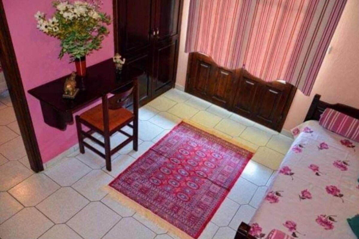 A bedroom with a bed, a chair, a desk, and a small table with a vase of flowers.