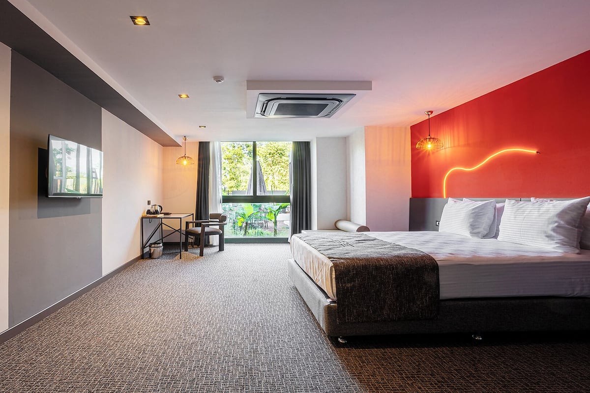A modern hotel room with a bed, a desk, and a view of greenery outside.