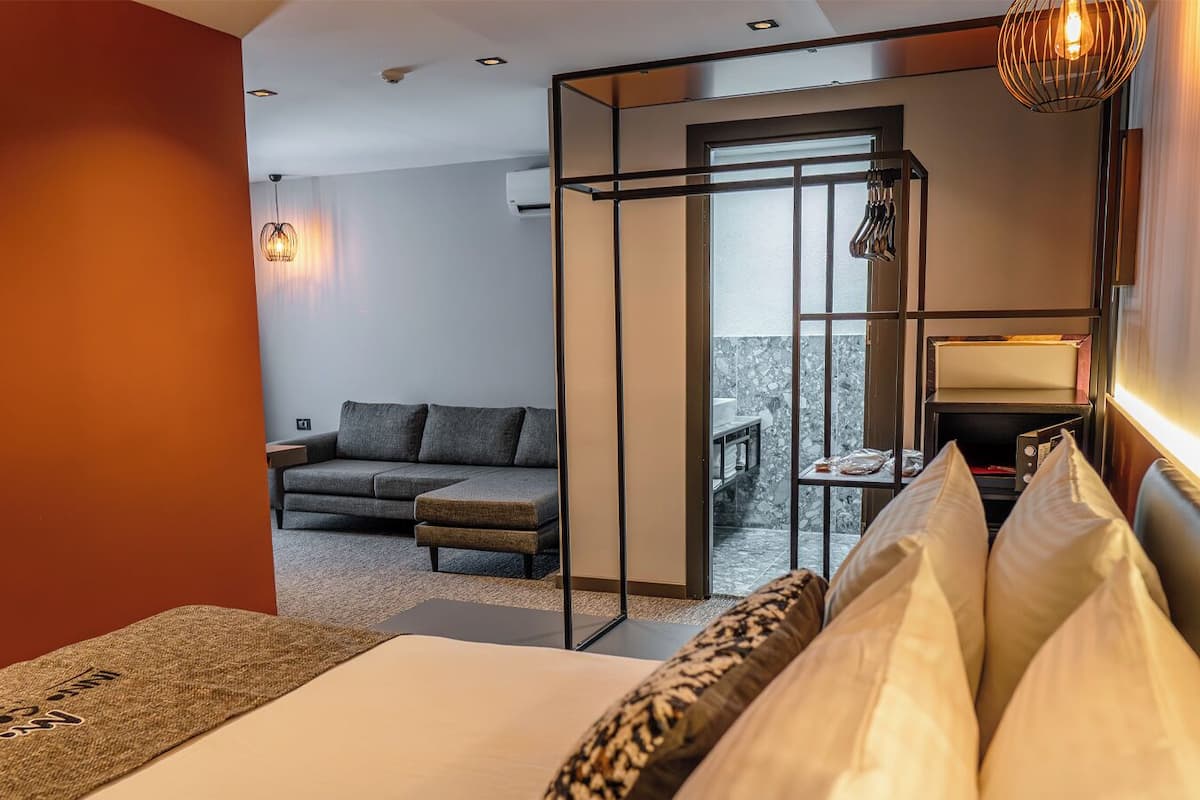 A hotel room with a bed, a sofa, a desk with a lamp, and a view of a balcony with a stone wall.