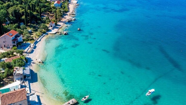 An aerial view of a coastal area with clear turquoise waters, a sandy beach, and buildings along the shore.